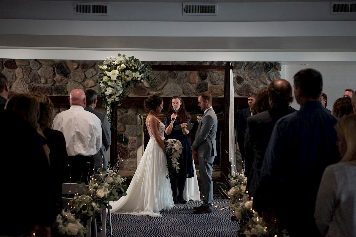 A wedding ceremony with a bride and groom facing each other, officiant in the middle, and guests seated on both sides in a decorated indoor venue.