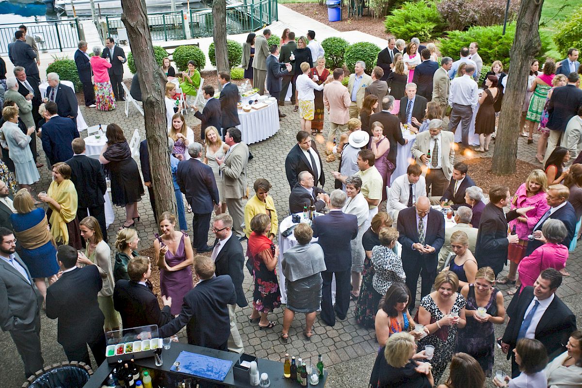 A large group of people are gathered at an outdoor event, mingling and socializing around tables and a drink station under trees.