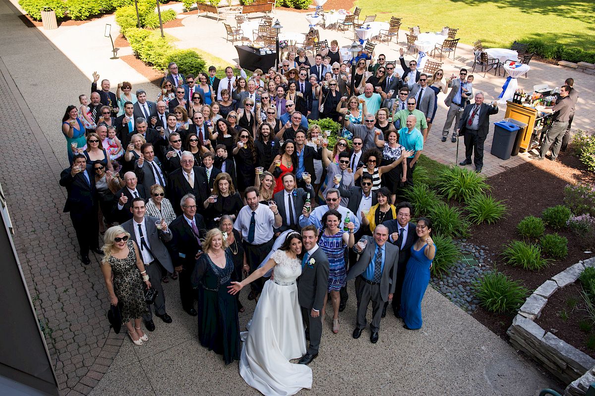 A large group of people, including a couple in wedding attire, are gathered outside for a group photo at what appears to be a wedding.
