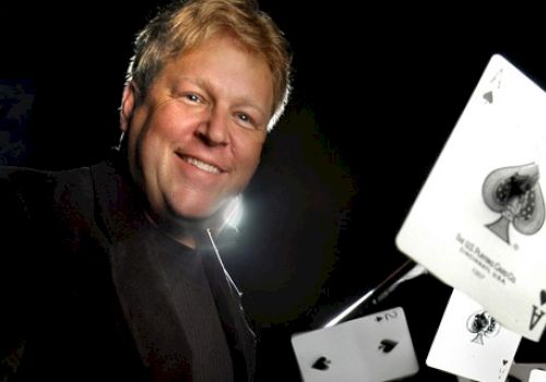 A person is smiling and appears to be performing a card trick, with several playing cards, including an ace of spades, floating in front of them.
