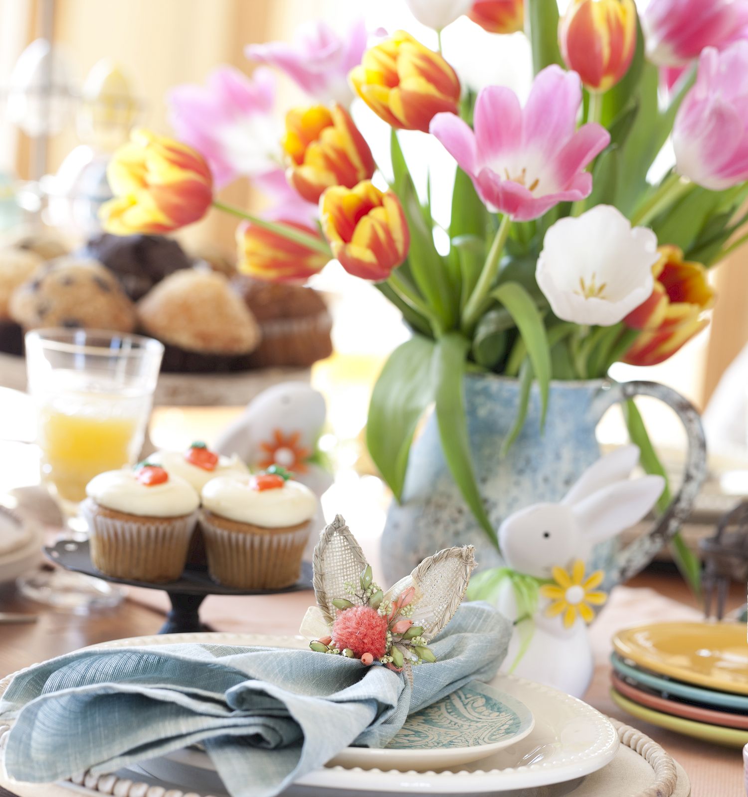A decorated table with flower arrangement, muffins, cupcakes, and juice. Pastel napkins, bunny figurines, and colorful tulips create a festive scene.