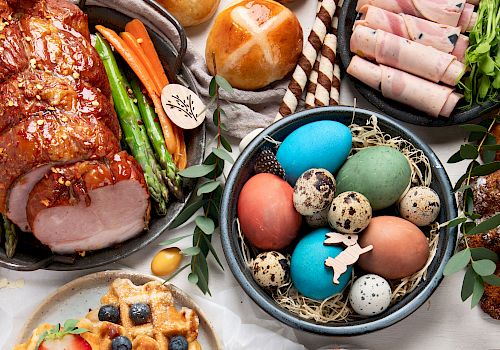 The image shows a festive spread including a glazed ham roast, colorful Easter eggs, hot cross buns, rolled deli meats, waffles, and vegetables.