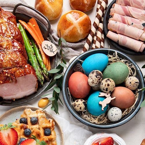 The image shows a festive spread including a glazed ham roast, colorful Easter eggs, hot cross buns, rolled deli meats, waffles, and vegetables.