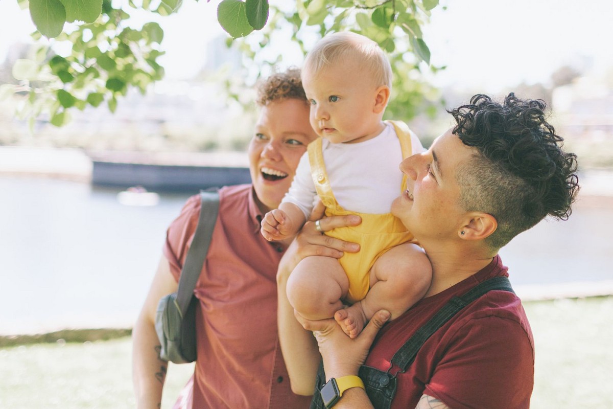 A happy group with two adults holding a baby under a tree by a river, enjoying a sunny day outdoors.