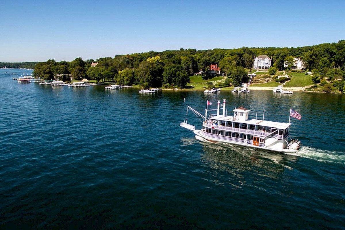 A boat sails on a large body of water near a shoreline with trees and houses; the sky is clear and blue, ending the sentence.