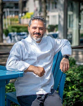 A person in a white chef's coat is sitting on a blue chair outdoors, smiling and resting their arm on a table, with greenery in the background.