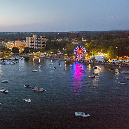 An aerial view of a waterfront with boats, a lit Ferris wheel, docks, and buildings in the evening. The skyline and horizon are visible.