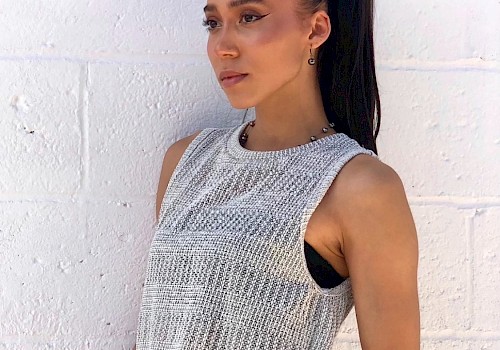 A person with a ponytail wearing a sleeveless knit top and matching pants, standing against a white brick wall, looking to the side.