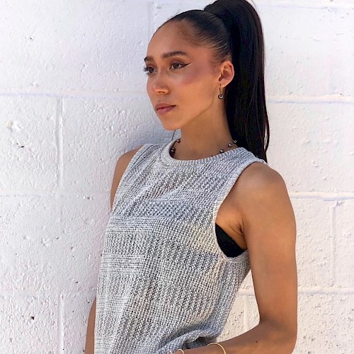 A person with a ponytail wearing a sleeveless knit top and matching pants, standing against a white brick wall, looking to the side.