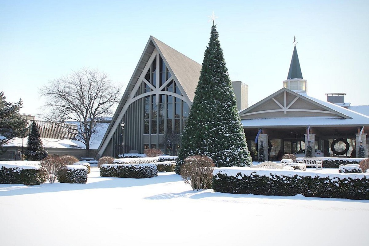 A snowy landscape featuring a church with a tall Christmas tree and evergreen shrubs under a clear blue sky, with snow covering the ground.