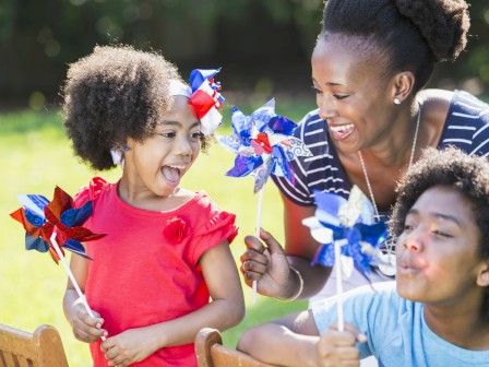 A woman and two children are enjoying a sunny day in a garden, playing with colorful pinwheels and smiling.