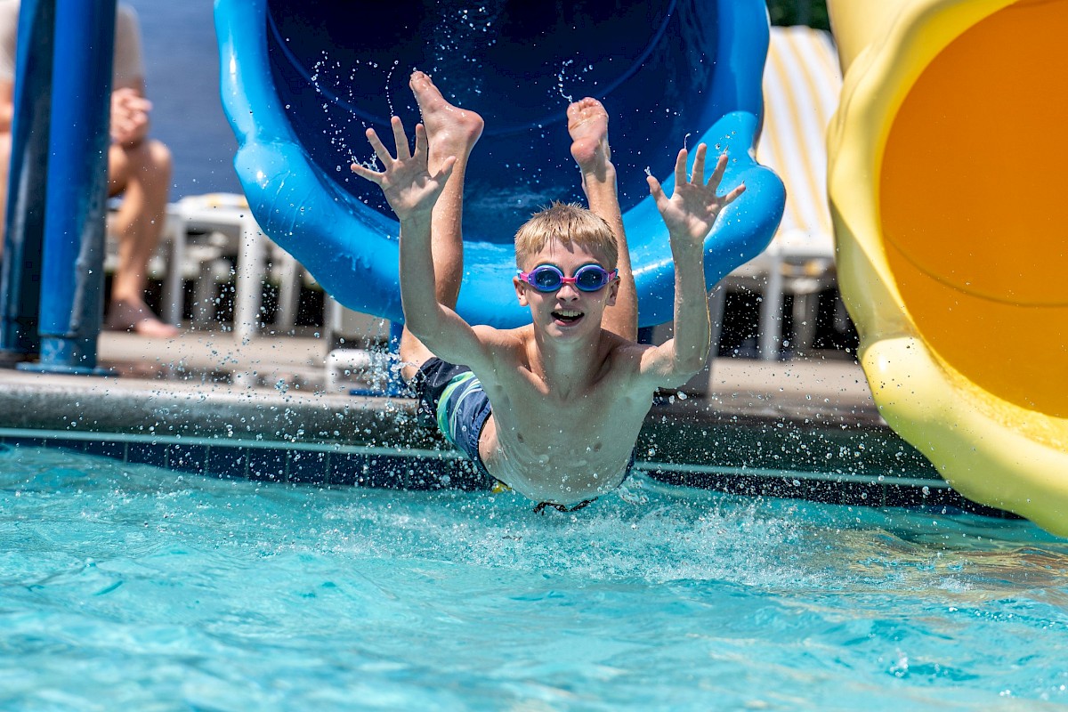 A boy wearing goggles is sliding out of a blue water slide into a swimming pool.