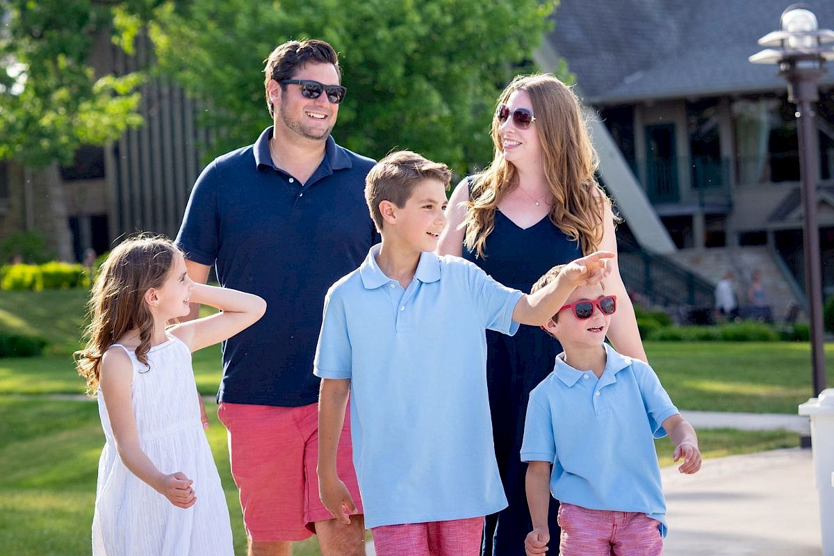 A family of five, including two children and two adults, are enjoying a sunny day outdoors near a modern building, smiling and interacting.
