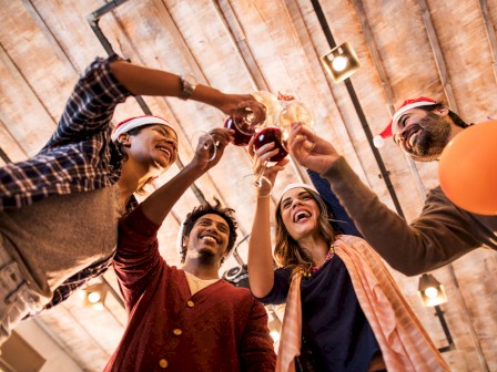 A group of people in festive attire raising their glasses in a celebratory toast, with balloons around them in a warmly lit setting.