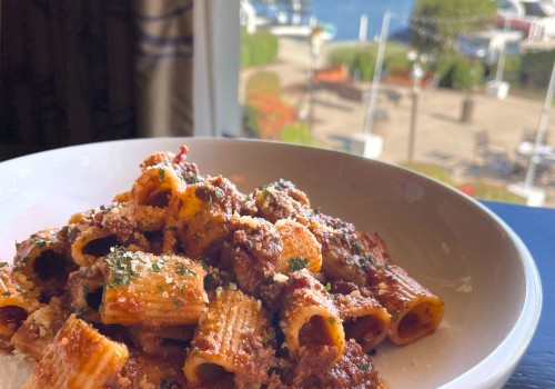 A bowl of rigatoni pasta with meat sauce sits on a table, with a scenic marina and boats visible through the window in the background.