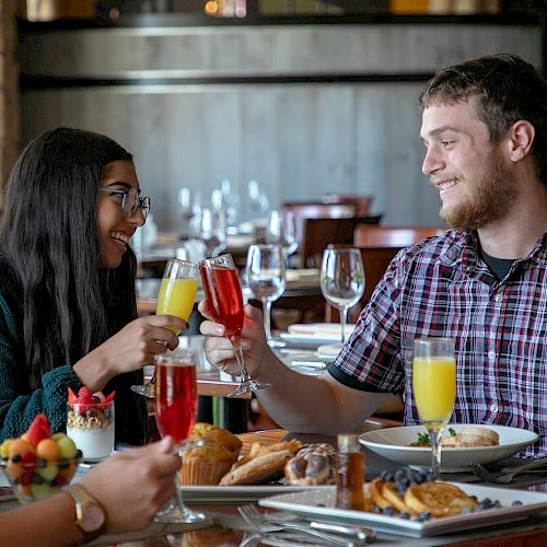 Two people are clinking glasses and smiling in a restaurant, with food spread out on the table, while two other people are visible.