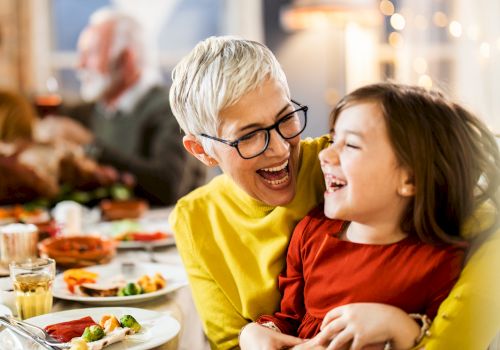 An older woman with glasses and short white hair laughs with a young girl in a red dress at a festive table. They appear to be enjoying a meal together.
