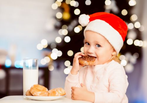 A child in a Santa hat enjoys a donut with milk beside a Christmas tree, glowing with lights in the background.