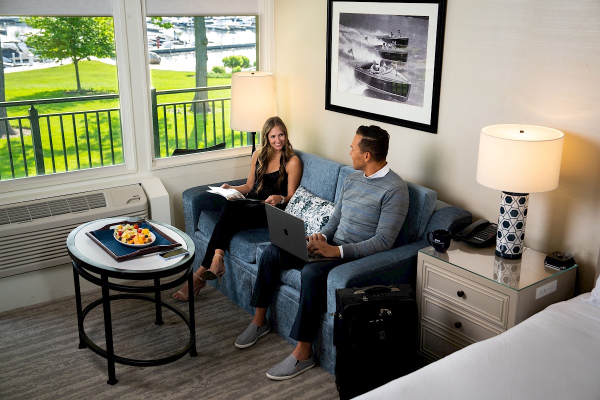 A man and woman sit on a sofa with laptops in a cozy room. A round table holds a fruit platter, and a window offers a scenic outdoor view.