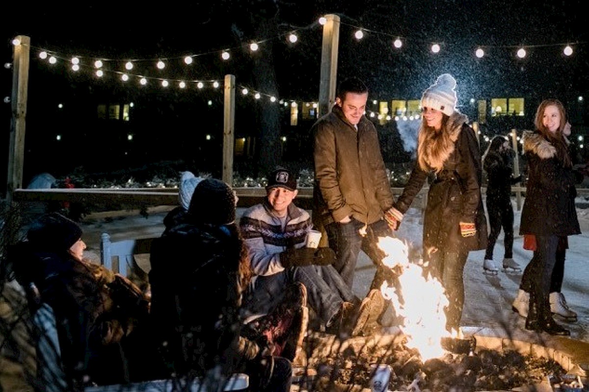 A group of people are gathered around a campfire under string lights on a snowy night, dressed warmly in coats and hats.