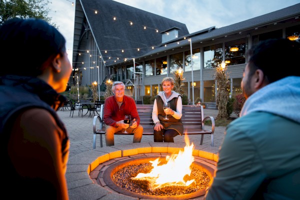 A group of people sitting around a firepit in front of a building with string lights, enjoying a casual evening outdoors.