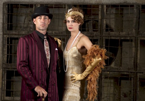 A man and woman in vintage 1920s attire pose in front of a rustic background, with the woman wearing a flapper dress and feathered accessories.