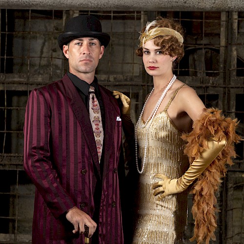 A man and woman in vintage 1920s attire pose in front of a rustic background, with the woman wearing a flapper dress and feathered accessories.