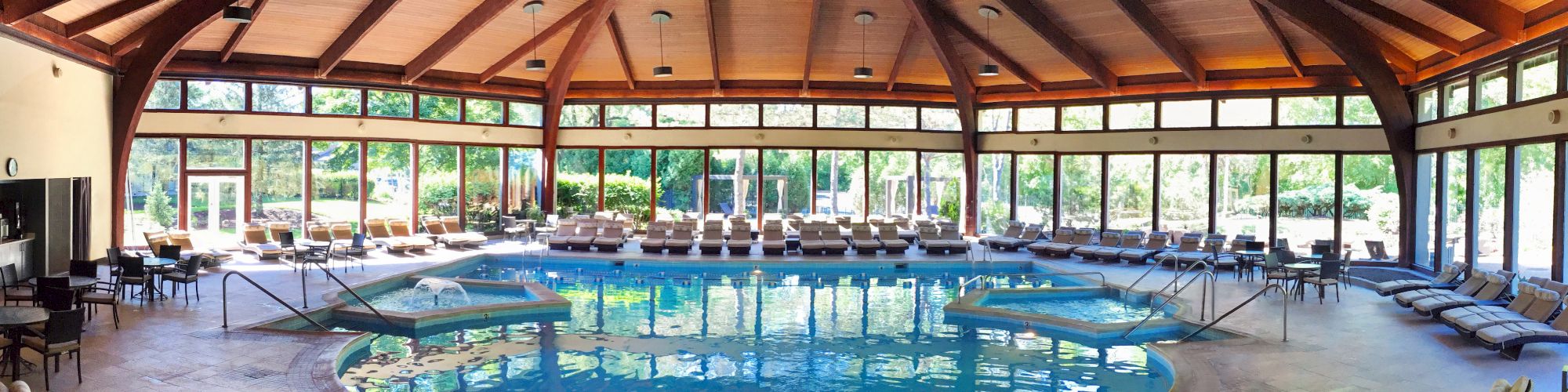 This image shows a spacious indoor pool area with a wooden ceiling, surrounded by lounge chairs and large windows offering a view of outdoor greenery.