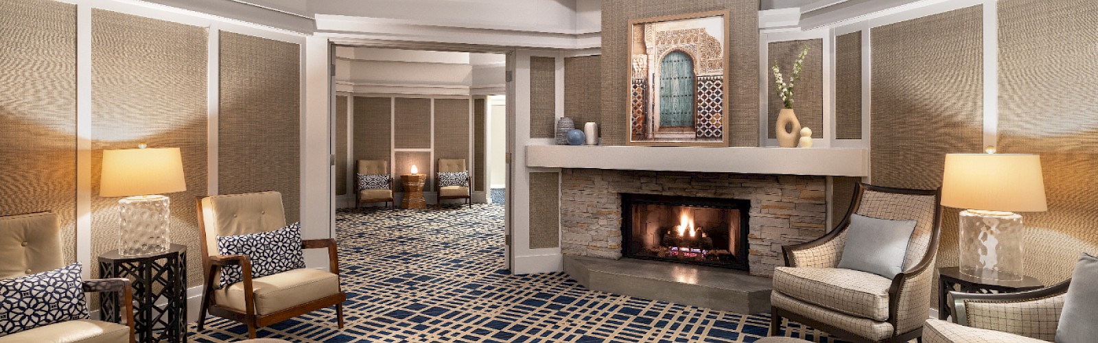 A luxurious spa lobby with a modern fireplace, comfortable seating in neutral tones, patterned carpet, and ambient lighting.
