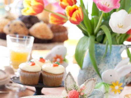 A table setting with a blue napkin, cupcakes, orange juice, muffins, and a vase of tulips, adorned with bunny-themed decorations.