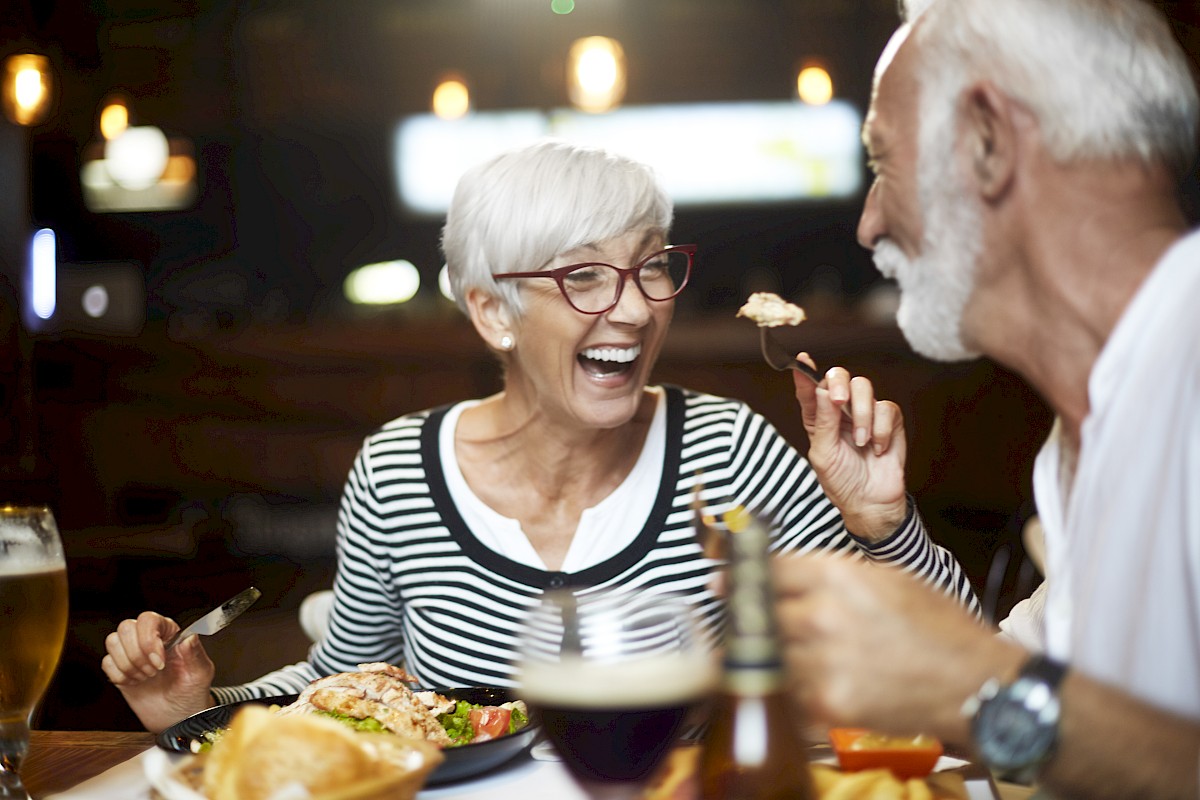 An older couple enjoys a lively meal together, laughing and eating at a restaurant with drinks and food on the table.