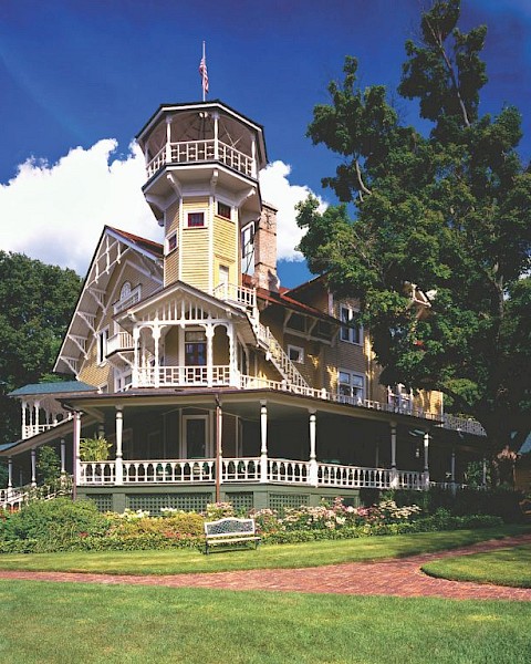 Victorian-style house with a tall tower, surrounded by greenery and a clear blue sky, featuring a charming wraparound porch.