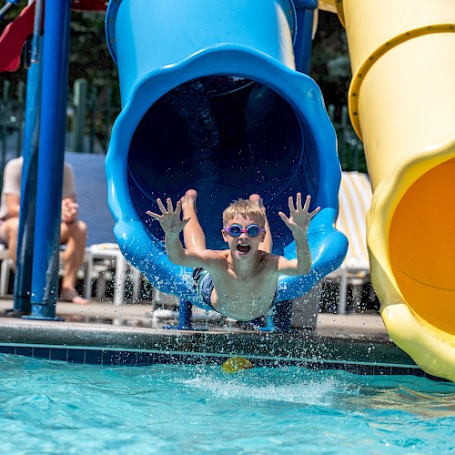 A child splashes out of a blue water slide into a pool, smiling and wearing goggles, with another yellow slide beside it.