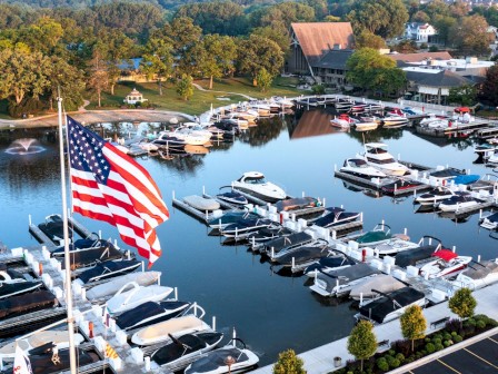 A marina with numerous boats docked, surrounded by trees and buildings, with an American flag prominently displayed in the foreground.