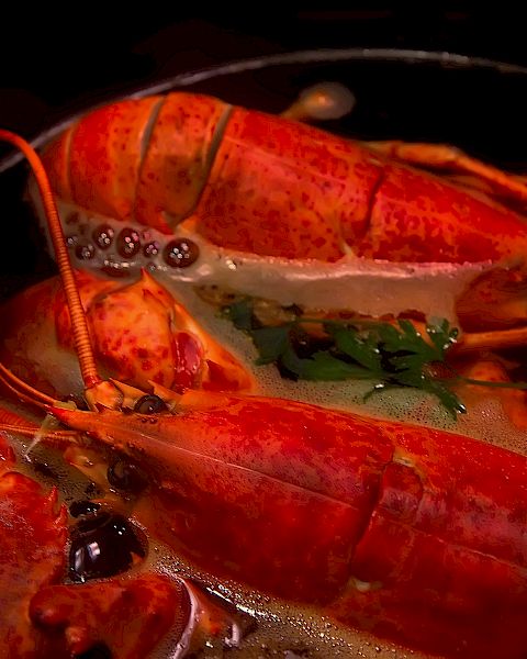 The image shows a dish with cooked lobsters in a sauce, garnished with herbs and served in a bowl.