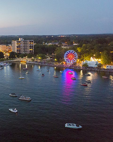 An aerial view of a waterfront with boats, a lit Ferris wheel, docks, and buildings in the evening. The skyline and horizon are visible.