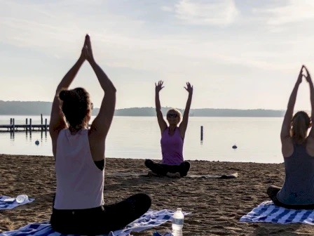 People are practicing yoga on a sandy beach by the water, with towels and water bottles nearby.