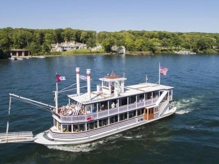A paddlewheel riverboat is cruising on a lake, surrounded by trees and houses, with passengers on deck.