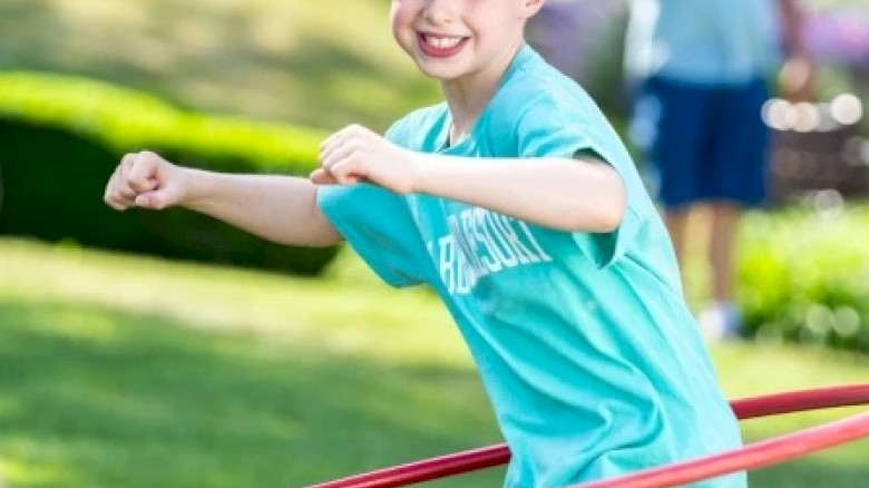 A child happily playing with a hula hoop in a sunny outdoor setting, wearing a blue shirt and smiling broadly at the camera.