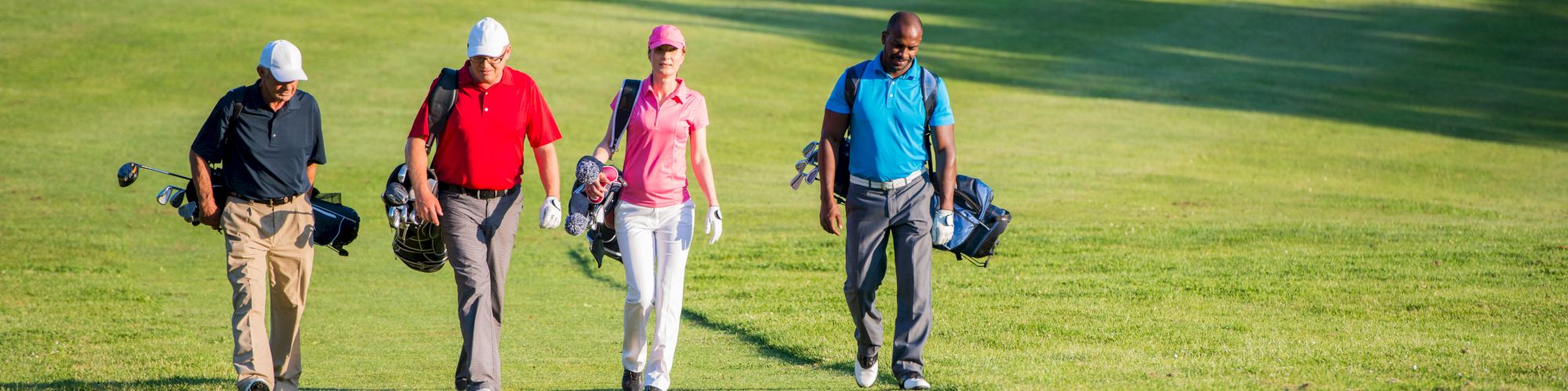 Four golfers are walking together on a golf course, carrying their bags, with a sand trap visible in the background.