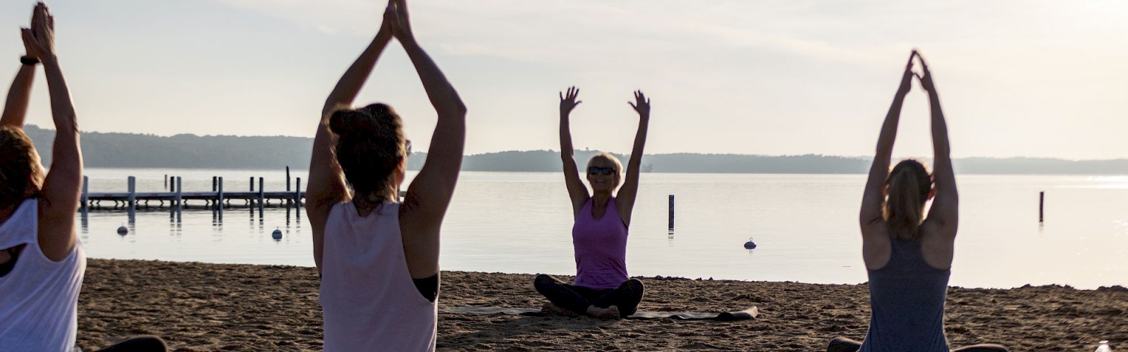A group of people doing yoga on a beach with mats, near a body of water, with a pier in the background and the sun setting or rising.