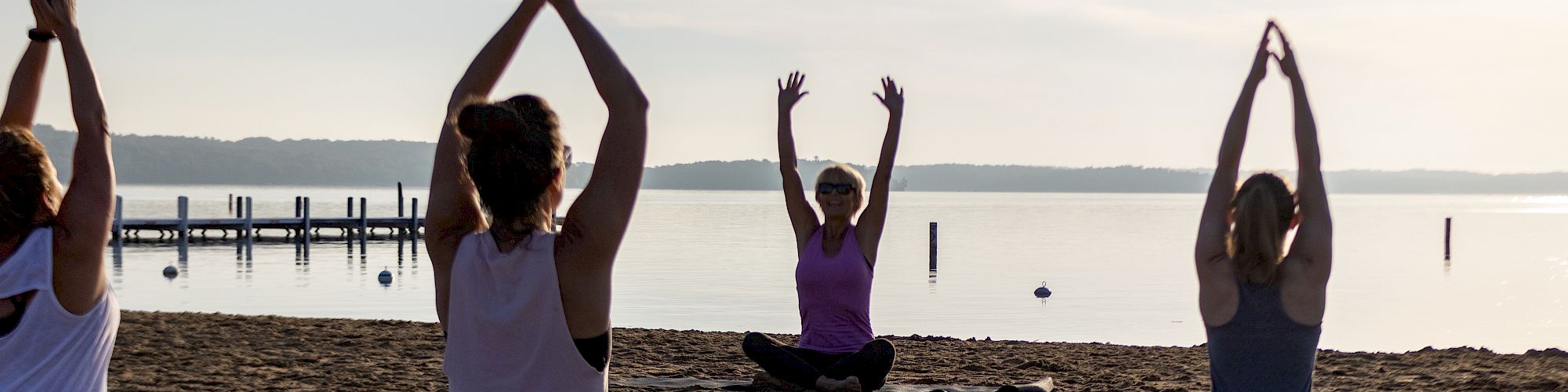 A group of people doing yoga on a beach with mats, near a body of water, with a pier in the background and the sun setting or rising.