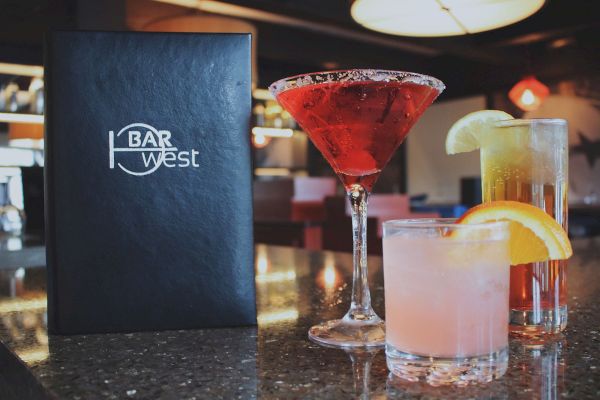 A Bar West menu is on a counter with three cocktails: a red martini, a lemon-garnished drink, and a pink drink with an orange slice.