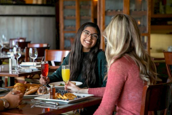 Two women sitting at a restaurant table, enjoying drinks and meals while having a conversation and smiling at each other.
