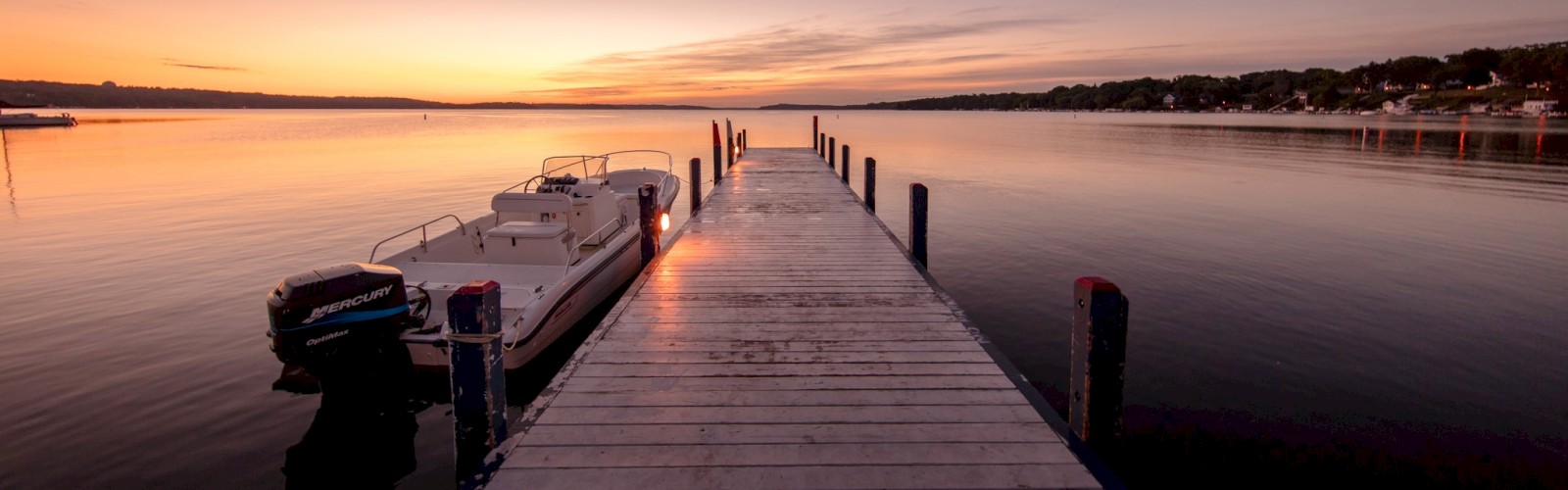 A scenic view from a wooden dock extending into calm water at sunset, with a moored boat to the left.