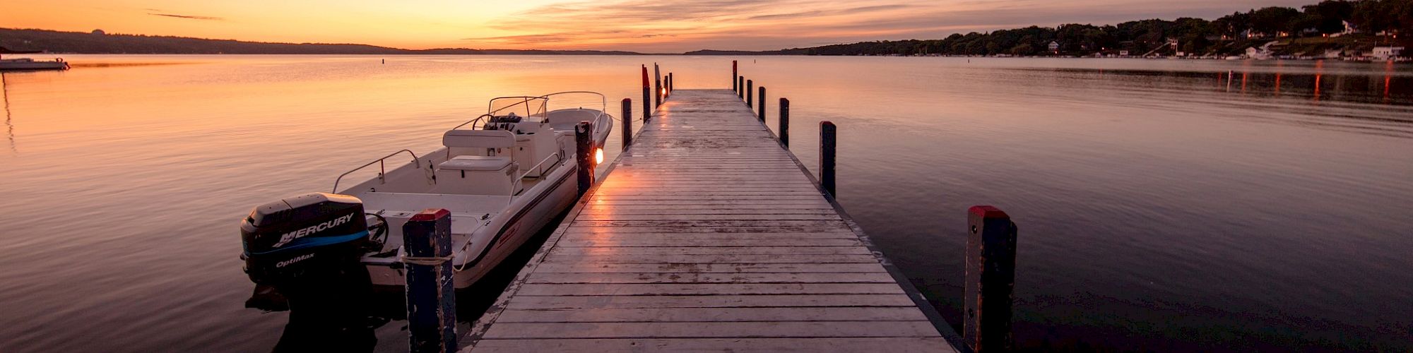 A scenic view from a wooden dock extending into calm water at sunset, with a moored boat to the left.