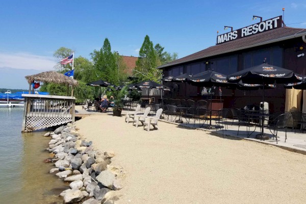 A lakeside resort with outdoor seating under umbrellas, a wooden dock, and a sign reading 