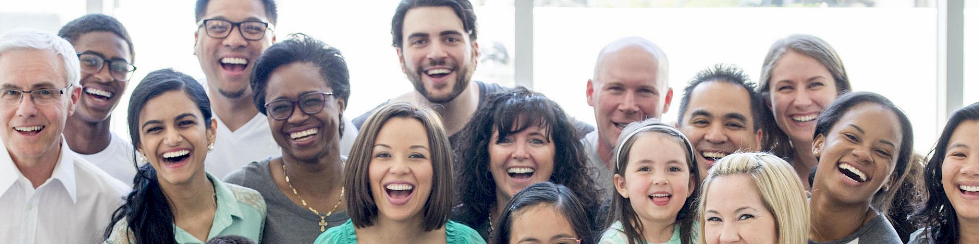 A diverse group of people, including men, women, and children, smiling and standing close together indoors.