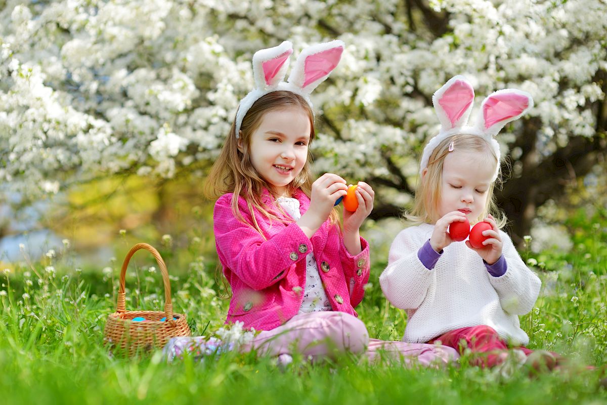 Two children wearing bunny ears sit on grass with a basket, holding colorful Easter eggs, against a background of blooming trees.