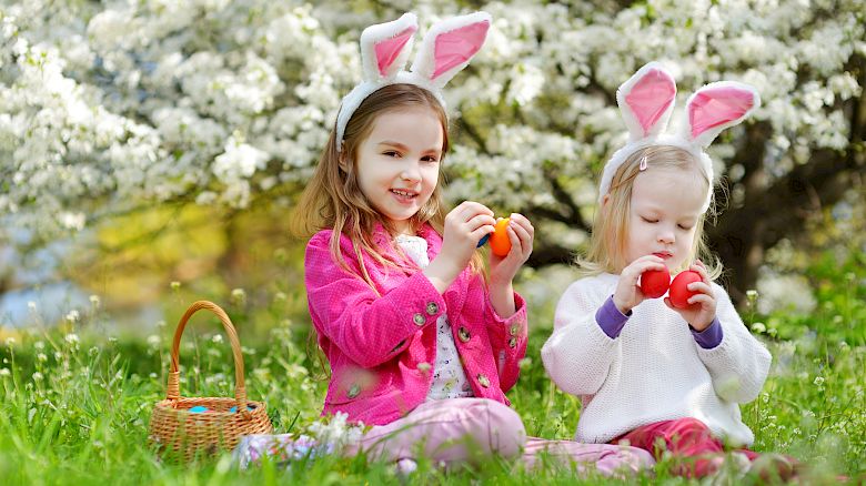 Two children wearing bunny ears sit on grass with a basket, holding colorful Easter eggs, against a background of blooming trees.